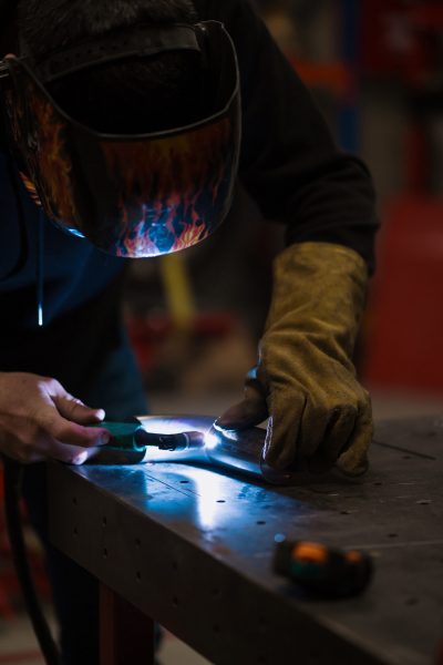 Worker in helmet with flames welding the steel part of a motorbike using an argon welding machine and gloves in a garage